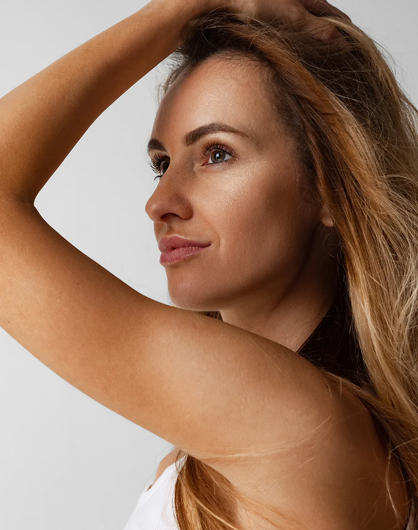 Close-up of a woman with light brown/blonde hair, her head and upper body angled slightly to her right. She gazes upward, her right hand gently resting on her hair. Her skin appears smooth and tanned, with a soft highlight on her cheekbones. Her expression is neutral yet contemplative, and her lips are a natural rosy pink. Her hair falls loosely around her shoulders against a plain, light gray background. - Brow Lift in Youngstown, OH