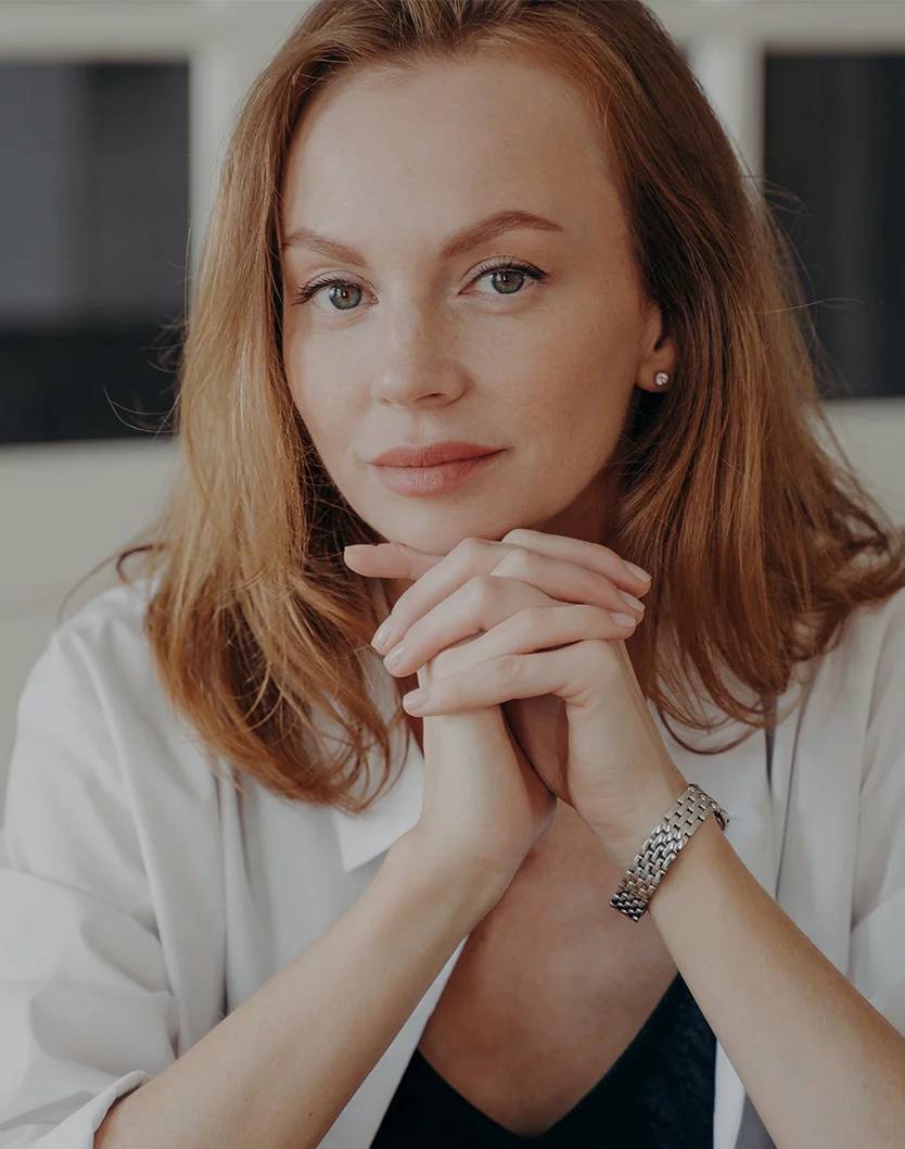 A woman with shoulder-length reddish-brown hair looks directly at the camera with a calm expression. Her hands are clasped under her chin. She wears a light-colored button-down shirt, a silver-toned bracelet on her left wrist, and a small stud earring. The softly lit, blurred background suggests an indoor setting. - Buccal Fat Removal in Youngstown, OH