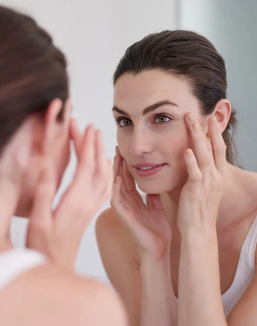 A light-skinned woman with dark brown hair in soft waves gazes at her reflection in a mirror, hands gently placed on her cheeks and jawline. Her expression is pleasant and focused. She wears a white sleeveless top, and the soft, even lighting highlights her smooth skin and hair. The light, neutral-toned background suggests a bathroom or vanity, keeping the focus on her and her reflection. - Chemical Peels in Youngstown, OH