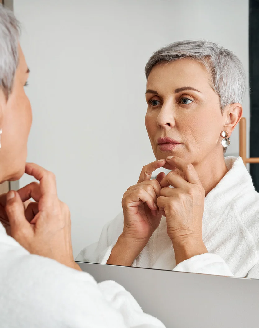 A middle-aged woman with short gray hair gazes at herself in a mirror, wearing a white bathrobe. Her hands gently touch her jawline. The lighting is soft and even, with a light gray background. The mirror reflects her and the blurred room behind, keeping the focus on her face and hands. - Chin Implant in Youngstown, OH