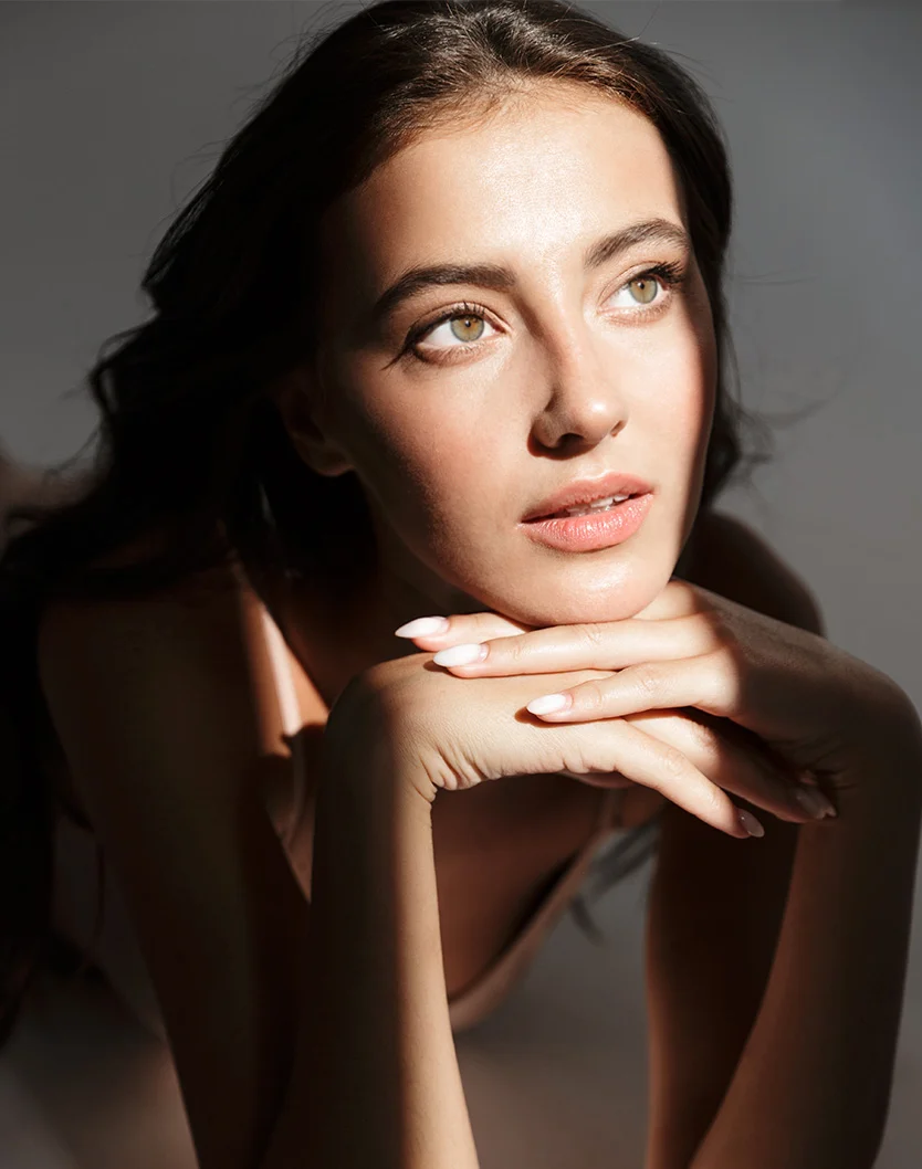 A close-up portrait of a woman with long, dark brown hair framing her face. Her light-toned skin has a natural glow, with soft lighting accentuating her cheekbones and delicate features. Her hazel eyes gaze off to the right, and her expression is calm and thoughtful. Resting her hands gently on her chin and cheeks, she wears light, french-tipped nails. The neutral gray background keeps the focus on her, enhancing the image’s elegant and sophisticated feel. - Co2 Laser Rejuvenation in Youngstown, OH