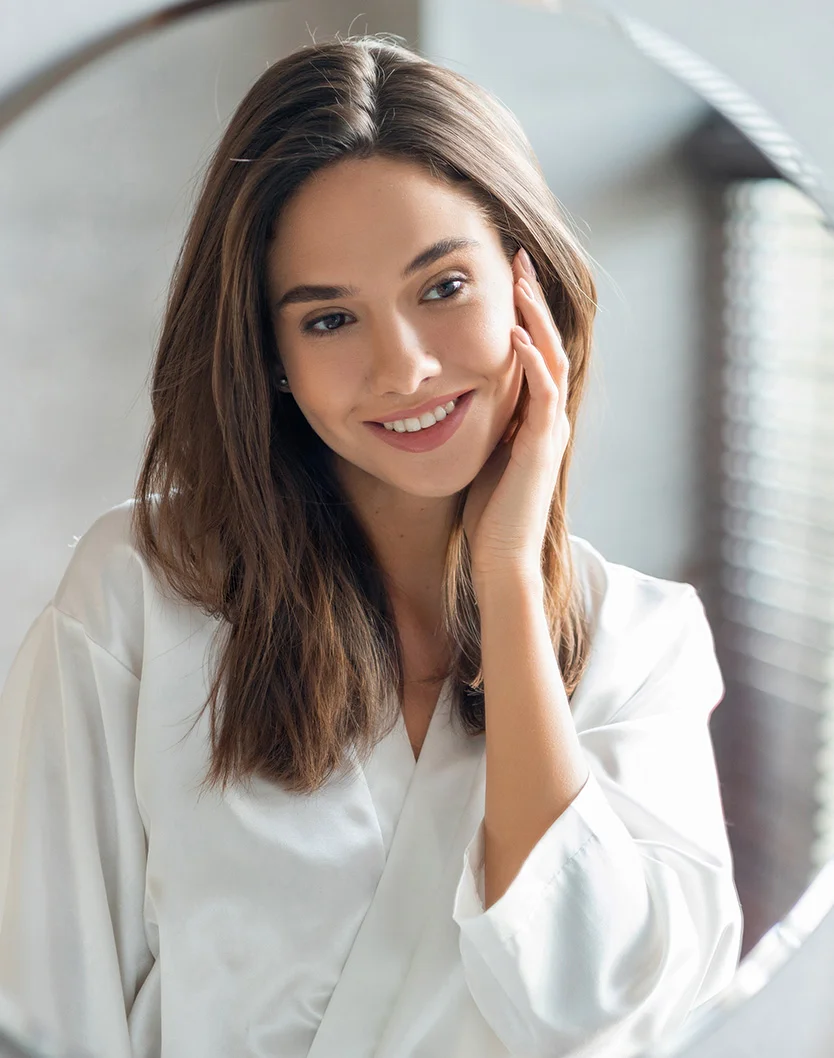 A young woman with long brown hair smiles while touching her cheek in a mirror. Her expression is pleasant and relaxed. She wears a white robe-like garment, likely satin, and the soft, natural lighting enhances the serene atmosphere. The blurred background suggests an interior space, with parts of a window and blinds or shades visible. The mirror is circular, reflecting the woman’s image. - Dermal Fillers in Youngstown, OH