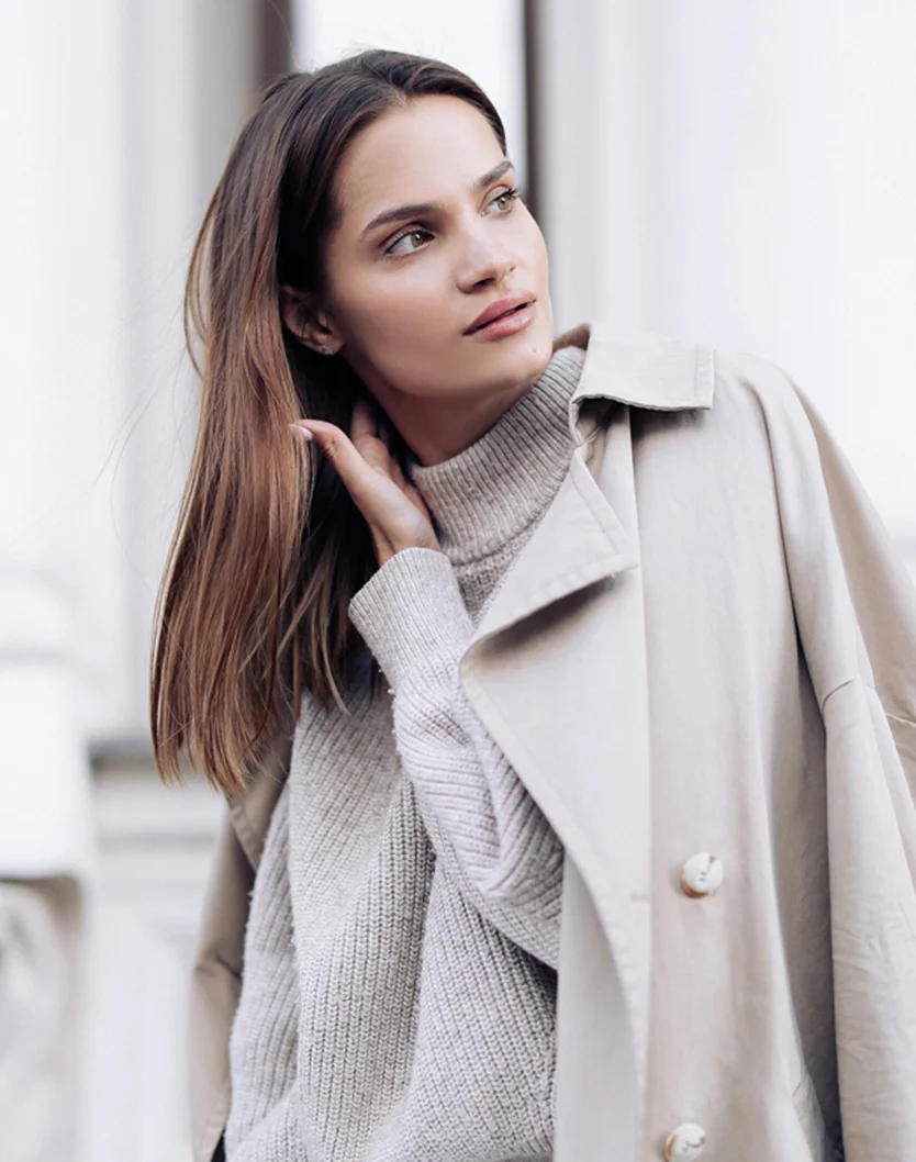 A woman with shoulder-length brown hair and a light complexion is pictured in a close-up. She looks off to her right, her expression thoughtful and relaxed. She wears a beige ribbed knit sweater with a turtle neck, layered under a beige trench coat with light-colored buttons. The background hints at an urban setting, with pale, muted architectural features softly blurred, complementing her outfit's light beige tones. - Exosomes for Hair Restoration in Youngstown, OH