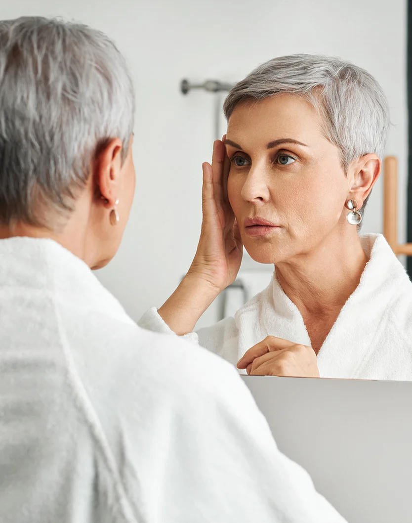 A middle-aged woman with short gray hair gazes at herself in a mirror, wearing a white bathrobe. Her hands gently touch her jawline. The lighting is soft and even, with a light gray background. The mirror reflects her and the blurred room behind, keeping the focus on her face and hands. - Exosomes in Youngstown, OH