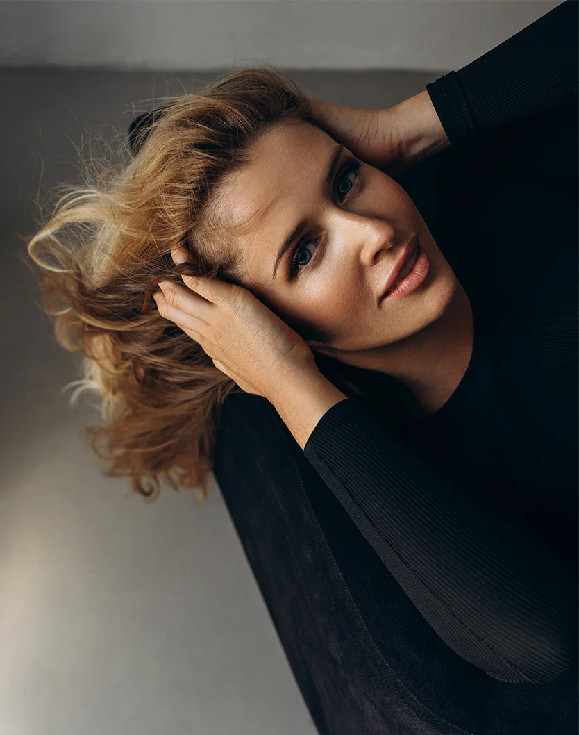 A high-angle, close-up of a woman with shoulder-length, light brown curly hair. Her hands are resting on either side of her temples, supporting her head. She has soft, relaxed expression, with light blue eyes looking directly forward and light pink lips. She wears a black, close-fitting long-sleeved top with a subtle ribbed texture. The background is a muted taupe or gray wall, with soft, diffused lighting casting no harsh shadows, focusing on her face and hair. - Eyelid Surgery in Youngstown, OH