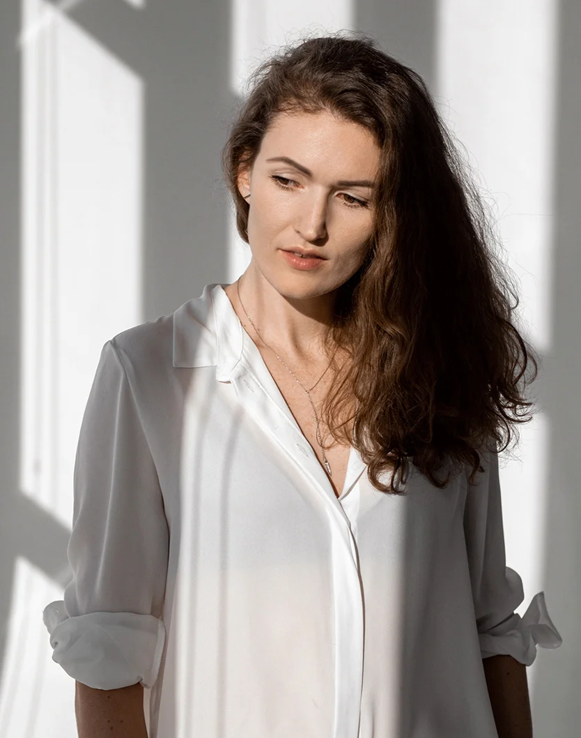A woman with long, wavy brown hair looks downward, wearing a loose white button-down shirt. The soft, diffused lighting and vertical shadows from a nearby window create a contemplative mood. - Facetite Acutite in Youngstown, OH