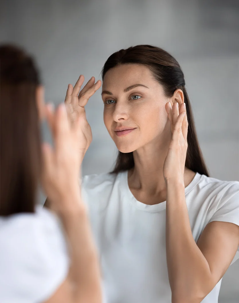 Close-up of a light-skinned, middle-aged woman with medium brown hair, looking at herself in a mirror. She wears a plain white short-sleeved t-shirt and touches her brow, cheek, and neck with her hands, her expression pleasant and attentive. The reflection in the mirror is slightly blurry, creating a soft and slightly distorted image. The background is a neutral light gray, out-of-focus, with soft, even lighting. - IPL Photorejuventation in Youngstown, OH