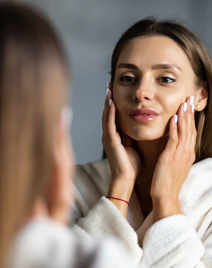 Close-up of a light-skinned woman with shoulder-length brown hair, wearing a light beige or cream-colored robe. She gently touches her face, specifically her jawline and the sides of her face, with a thoughtful and serene expression. Her lips have a natural rosy tint, and a thin red bracelet is visible on her wrist. The background is a muted gray, out-of-focus, creating an intimate feel. A blurred reflection of the woman in the mirror adds depth to the image, with the reflection slightly out of focus, enhancing the overall soft atmosphere. - Lasemd Ultra in Youngstown, OH