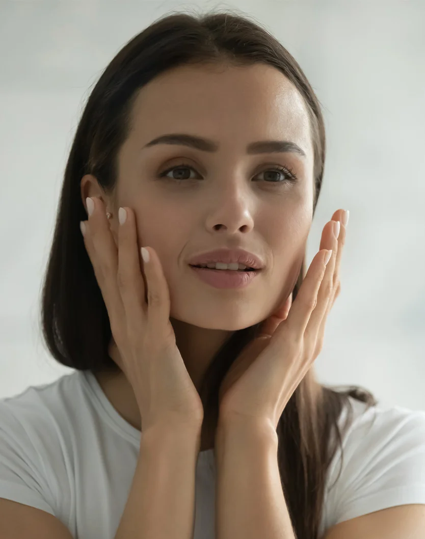 Close-up of a light-skinned woman with long, dark brown hair applying skincare. She has a gentle expression, with her hands lightly touching her cheeks and jawline, possibly applying cream or lotion. Her smooth, healthy skin and natural pink lips are highlighted, and she's wearing a plain white short-sleeved t-shirt. The soft, even lighting and neutral, blurred background focus attention on her face and hands. - Micro Laser Peel Erbium in Youngstown, OH