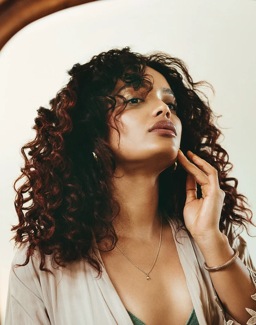 Close-up of a woman with curly dark brown hair, looking at her reflection. She wears a beige robe and light teal top, with a silver necklace and gold bracelet. Her expression is thoughtful, and the background is soft beige with a curved reflection surface. - Microneedling Exceed in Youngstown, OH