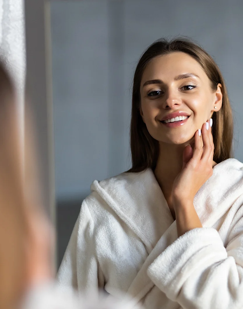 A young woman smiles while looking at herself in a mirror, gently touching her cheek. She wears a soft, plush white bathrobe with a hood. The background is a muted gray, and the reflection of the woman and her surroundings is slightly out of focus, adding depth to the image. - Necklift in Youngstown, OH