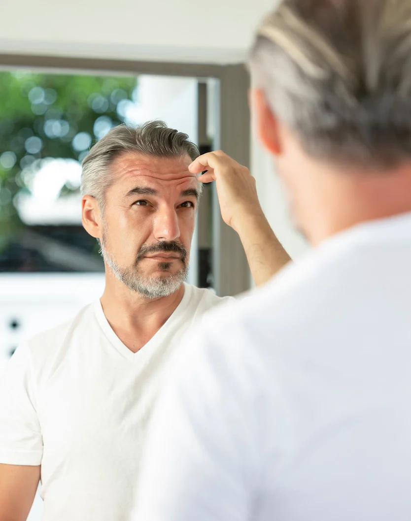 A middle-aged man with short gray hair and a trimmed beard looks at his reflection in a mirror. He has a thoughtful expression and is touching his forehead. He wears a plain white V-neck T-shirt. The background is slightly out of focus, suggesting an interior space with a window. His reflection shows his back, with the same hair and clothing. - Neograft in Youngstown, OH