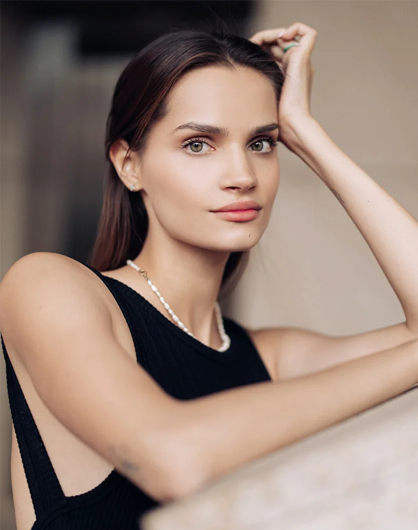 Close-up of a young woman with long dark brown hair and light-toned skin. She has light brownish-green eyes and a serene expression. She is wearing a black sleeveless top and a pearl necklace, with one hand resting on her head and the other on a beige surface. The background is a soft beige tone. - Otoplasty in Youngstown, OH