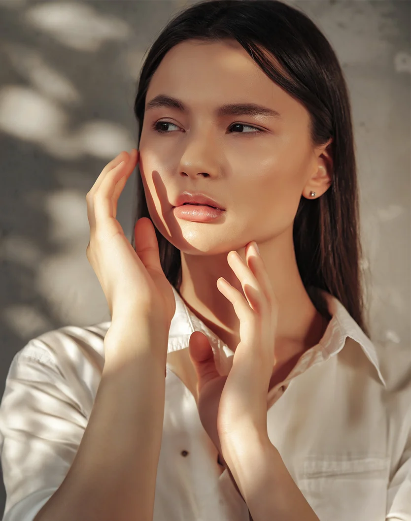 Close-up of a young woman with long, dark hair and smooth, light-toned skin. She has a gentle, contemplative expression. She wears a light cream-colored button-down shirt, possibly linen or cotton, with her hands gently caressing her face and neck. Soft, warm shadows fall across her skin and clothes, highlighting texture. The background features a light gray concrete or plaster wall, with patterns of light and shadow resembling leaves or branches, creating an intimate, feminine portrait. - Prescription Skincare in Youngstown, OH