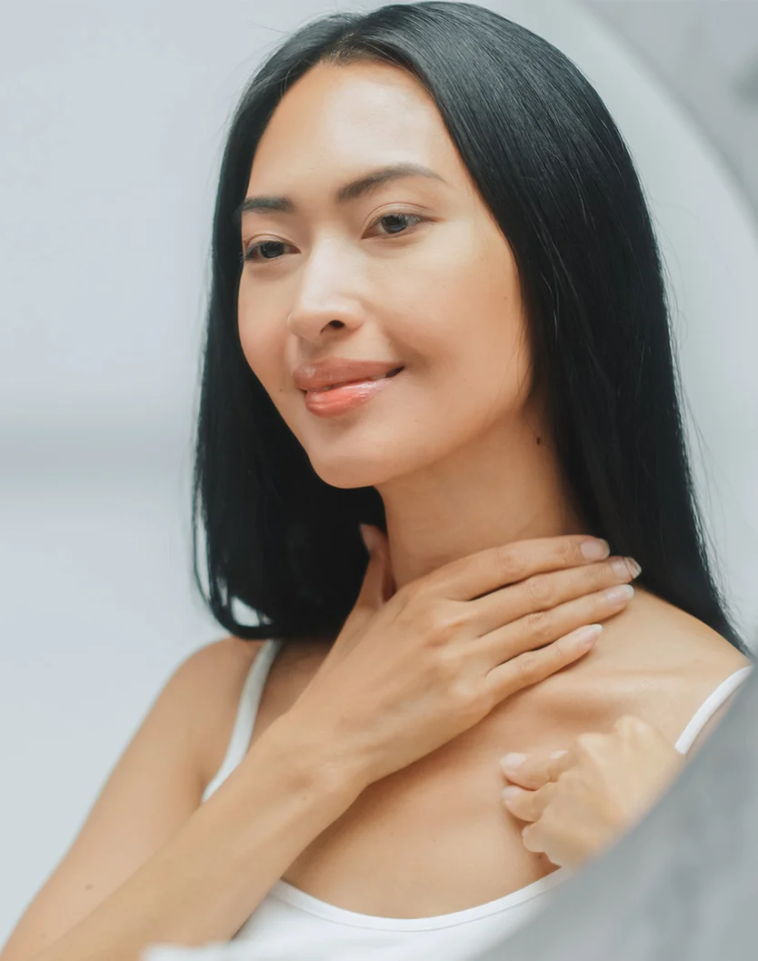 A light-skinned woman with long dark hair smiles gently while looking at herself in a mirror. She is wearing a white tank top and touches her neck and upper chest with her hands. The background is light gray-beige, with soft, diffused lighting highlighting her serene expression. - Renuvion Neck Lift in Youngstown, OH