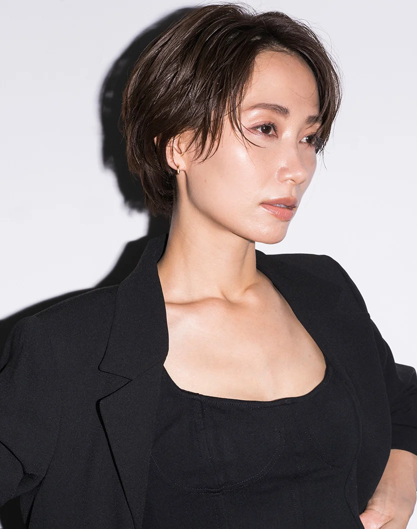Close-up of a woman with short, dark brown hair in a choppy bob. She is wearing a black blazer over a black fitted top, with subtle makeup and a small gold hoop earring. Her expression is neutral as she looks to her right. The background is plain white, with soft lighting highlighting her face and upper body. - Revision Rhinoplasty in Youngstown, OH