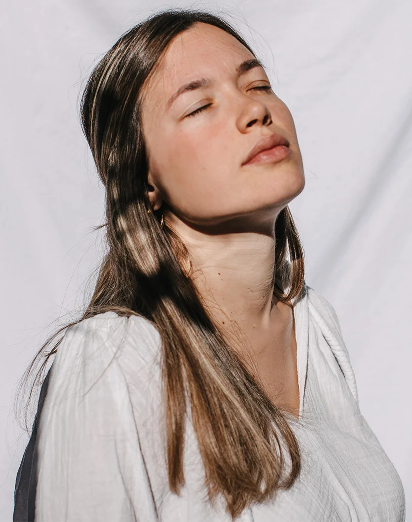Close-up of a young woman with light brown, shoulder-length hair. She has her eyes closed and a relaxed expression, wearing an off-white V-neck top. Her skin has a subtle rosy hue, and her lips are light pink. The background is a soft, pale off-white, with gentle shadows highlighting her face and shoulders. - Rhinoplasty in Youngstown, OH