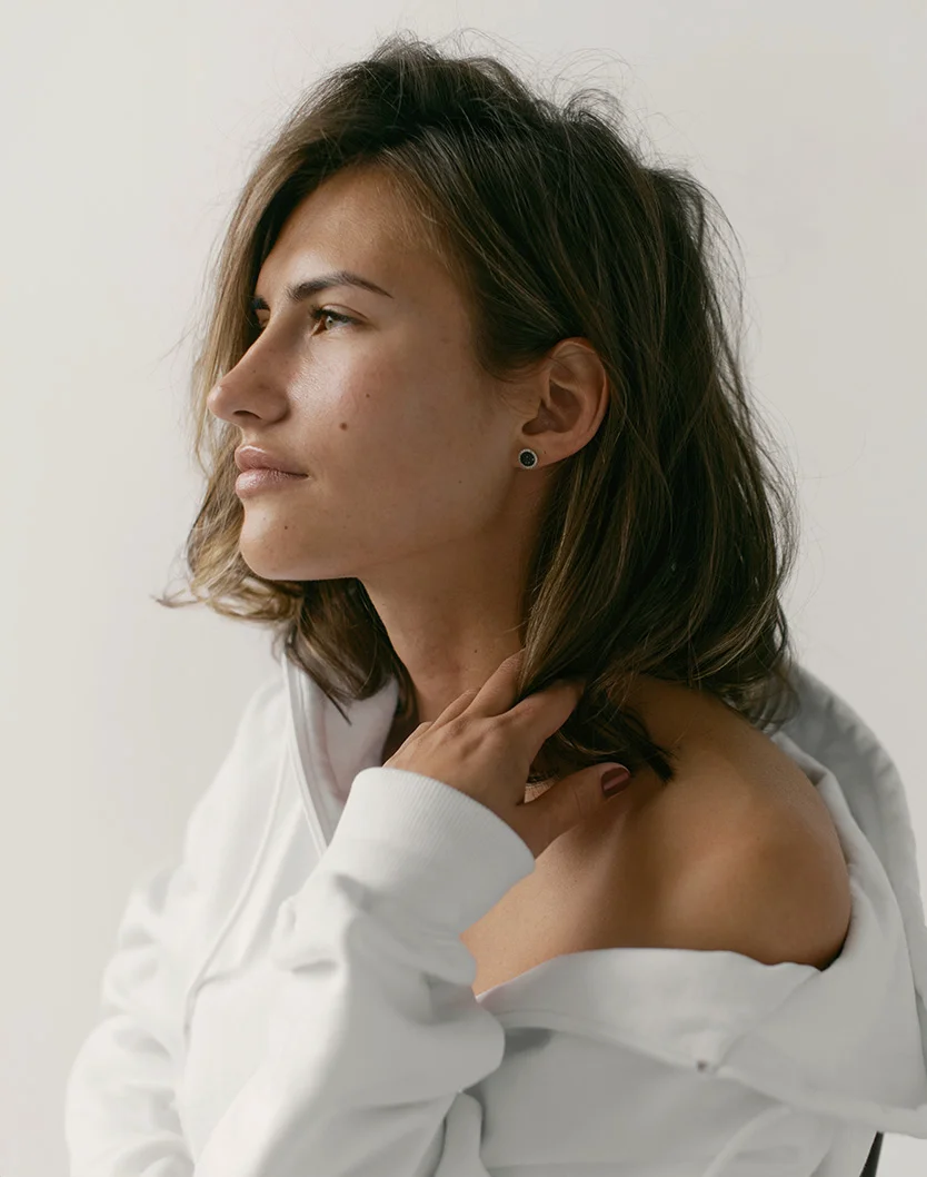 A light-skinned woman with shoulder-length, wavy brown hair looks slightly off to the viewer's right with a neutral expression. She wears a loose-fitting white top, and her hands rest gently on her neck and upper shoulder. Small, dark stud earrings are visible. The plain, light gray or white background provides a soft, minimalist contrast. - Therapeutic Facials in Youngstown, OH