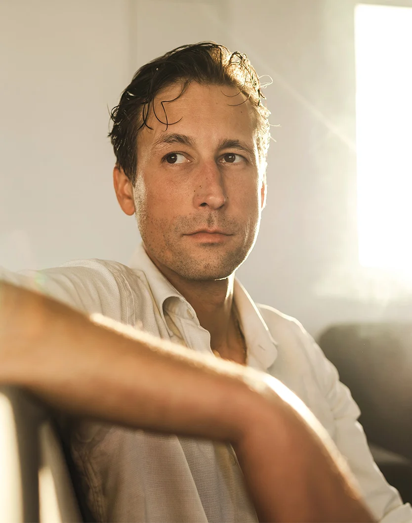 A light-skinned man in his 20s or 30s gazes slightly to his right with a neutral, contemplative expression. His dark, tousled hair appears damp or styled with product. He wears a beige button-up shirt, softly lit by what seems to be natural light. The blurred, pale grayish-white background suggests a nearby window or light source, creating a subtle glow around his face and shoulders. - Topical Treatments in Youngstown, OH