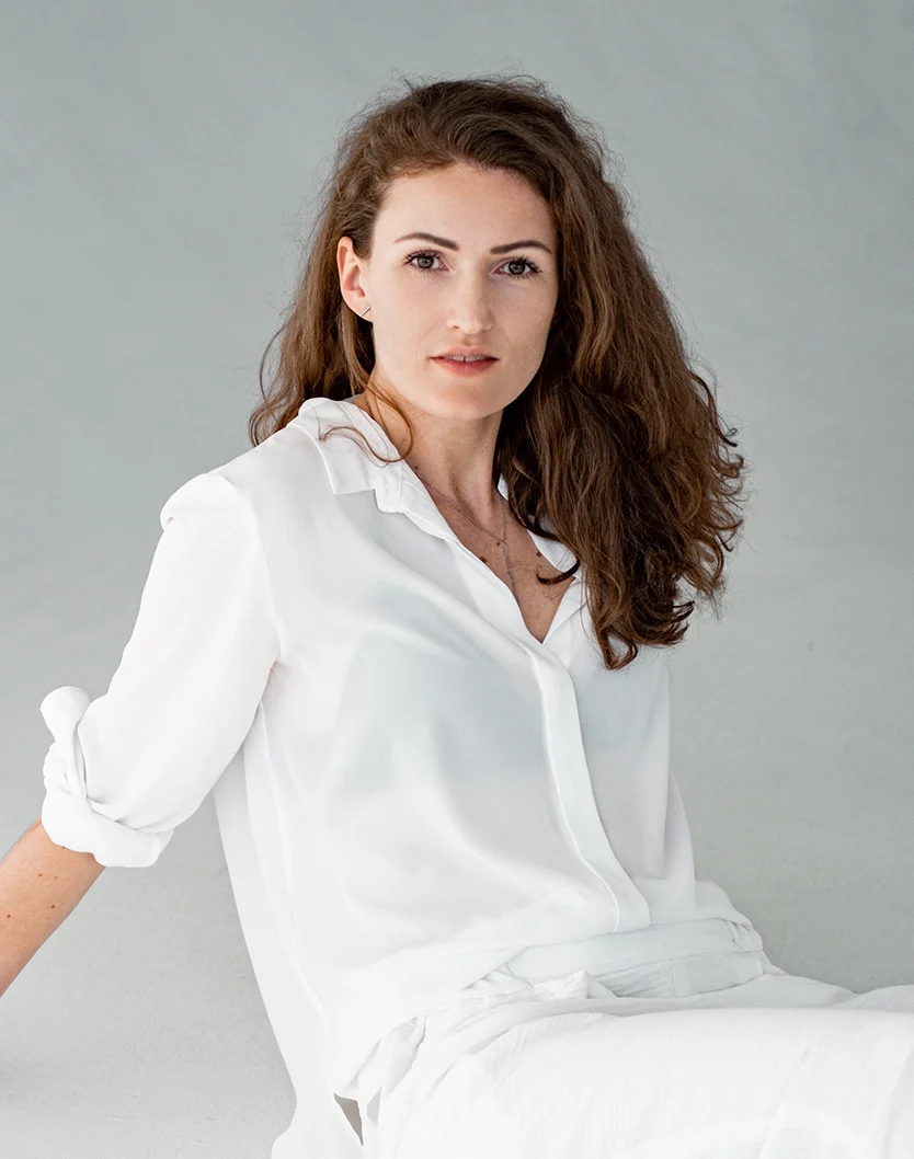 A medium shot of a light-skinned woman with shoulder-length, wavy brown hair. She wears a loose-fitting, white button-down shirt made of a smooth, slightly sheer material. Her expression is neutral, with her gaze directed at the viewer. The background is plain light gray, with even lighting and no harsh shadows. She is seated, with part of her lower body visible, and a necklace adorns her neck. - Vascular Birthmark Treatment in Youngstown, OH