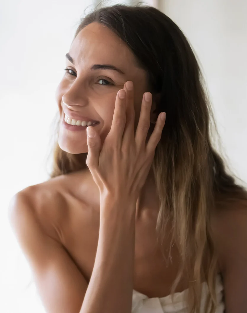 Close-up of a light-skinned woman with long, wavy, light brown hair applying skincare to her cheek. She is smiling with a pleasant, approachable expression. Wearing a white towel or robe, her smooth skin is highlighted against a soft, light gray/white background. - Venus Viva in Youngstown, OH